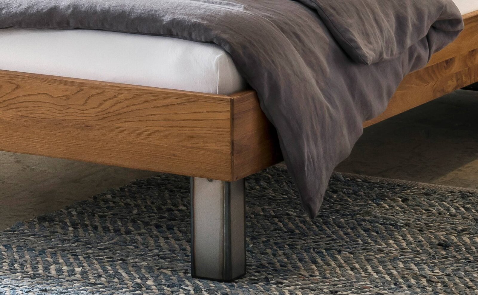 Detailaufnahme eines modernen Bettrahmens aus Holz mit Metallfuß, grauer Bettdecke und sichtbarer Matratze auf Teppich – Perspektive: seitliche Nahaufnahme am Fußende.