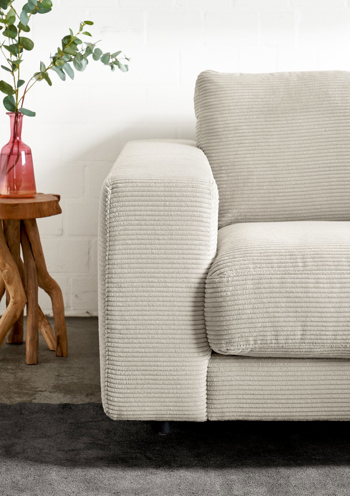 Nahaufnahme einer cremefarbenen Longchairkombination mit geripptem Stoffbezug, seitliche Perspektive. Daneben steht ein kleiner Holztisch mit einer roten Vase und Eukalyptuszweigen.