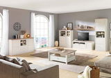 Helles Wohnzimmer mit einem weißen Sideboard im Landhausstil, frontal betrachtet, umgeben von weiteren passenden Möbeln und großen Fenstern.