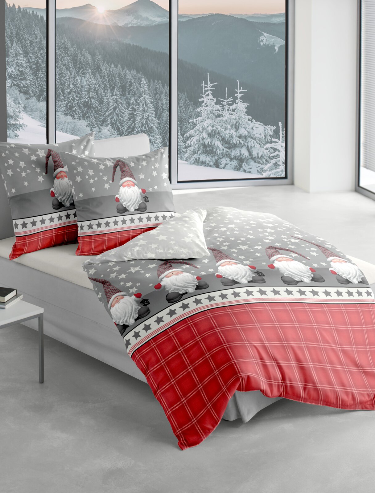 Bettwäsche-Set mit weihnachtlichem Design, bestehend aus grauen und roten Elementen mit Sternen und Weihnachtsmann-Motiven, auf einem Bett in einem modernen Raum mit Blick auf verschneite Berge.