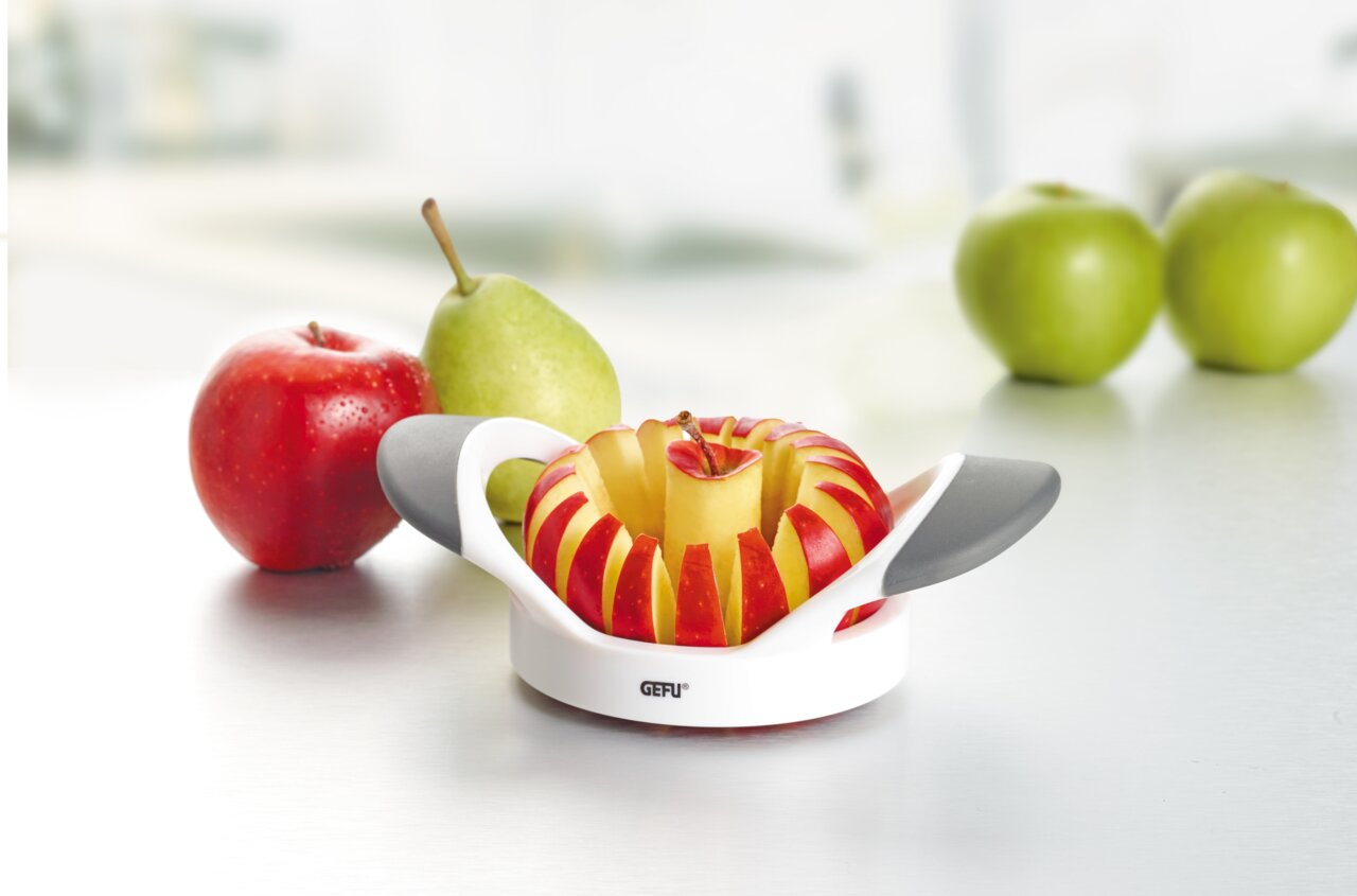 Apfelteiler Parti auf einer Küchenoberfläche, Vorderansicht, mit geschnittenem Apfel und weiteren Früchten im Hintergrund.