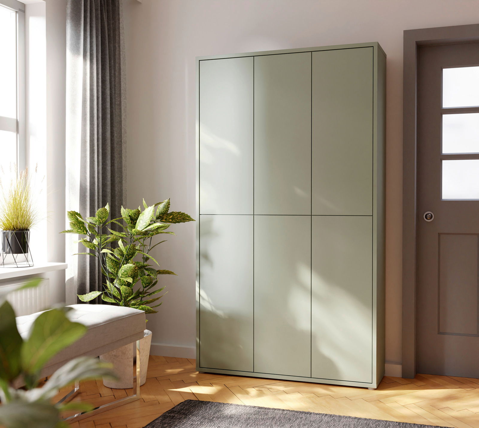Moderner, hoher XXL-Schuhschrank in einem hellen Raum, aus einer seitlichen Perspektive fotografiert. Der Schrank hat eine glatte, grüne Oberfläche und steht neben einer Tür und einem Fenster mit Pflanzen im Vordergrund.
