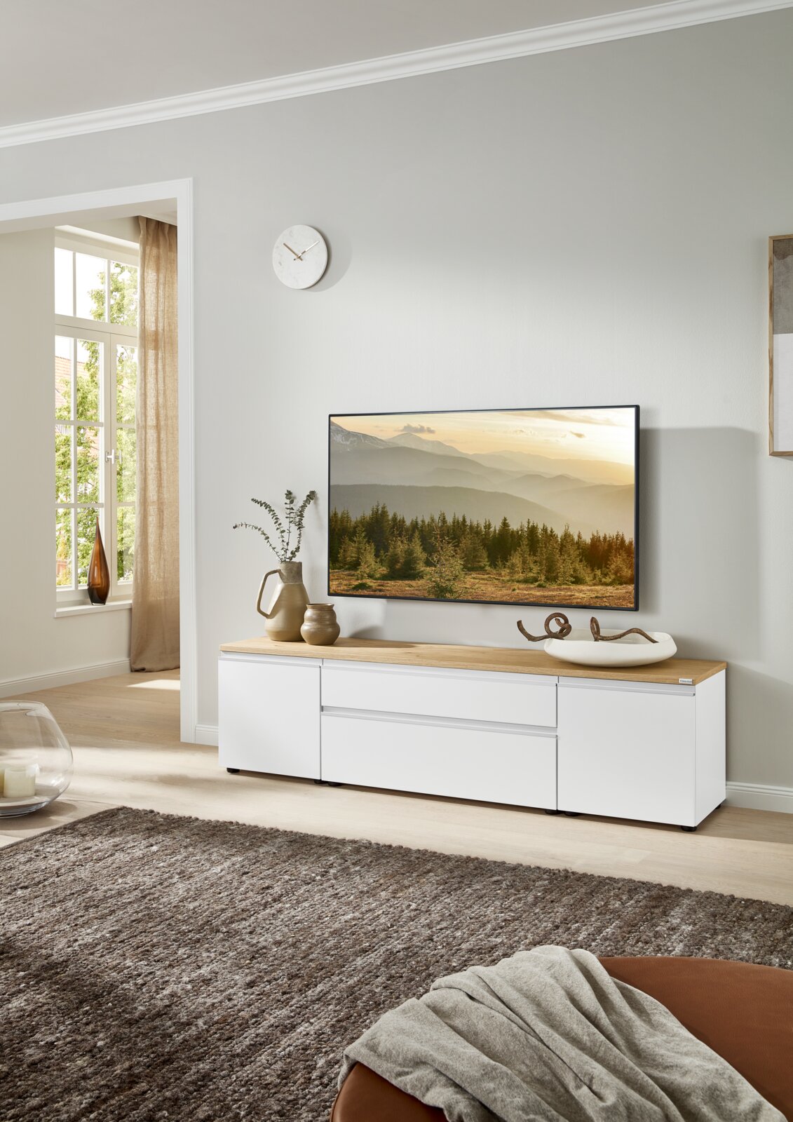 Modernes weißes TV-Lowboard mit Holzplatte und Schubladen im Wohnzimmer, frontal aus leicht schräger Perspektive fotografiert, mit Fernseher und Deko auf der Oberfläche.