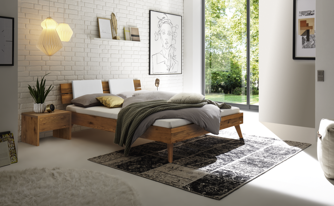 Modernes Schlafzimmer mit einem Kopfteil aus Holz und Metall, seitliche Perspektive. Das Bett ist mit weißen Kissen und einer grauen Decke dekoriert, umgeben von stilvollen Möbeln und Dekorationen.