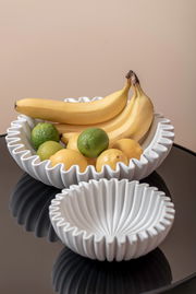 Zwei weiße, geriffelte Schalen auf einem Tisch, gefüllt mit Bananen, Limetten und Zitronen. Die Perspektive ist von oben.