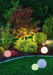 Verschiedene farbige Solar-Außenleuchten in Kugelform, die in einem Garten bei Nacht leuchten. Perspektive: seitlich auf die Leuchten und den Garten gerichtet.