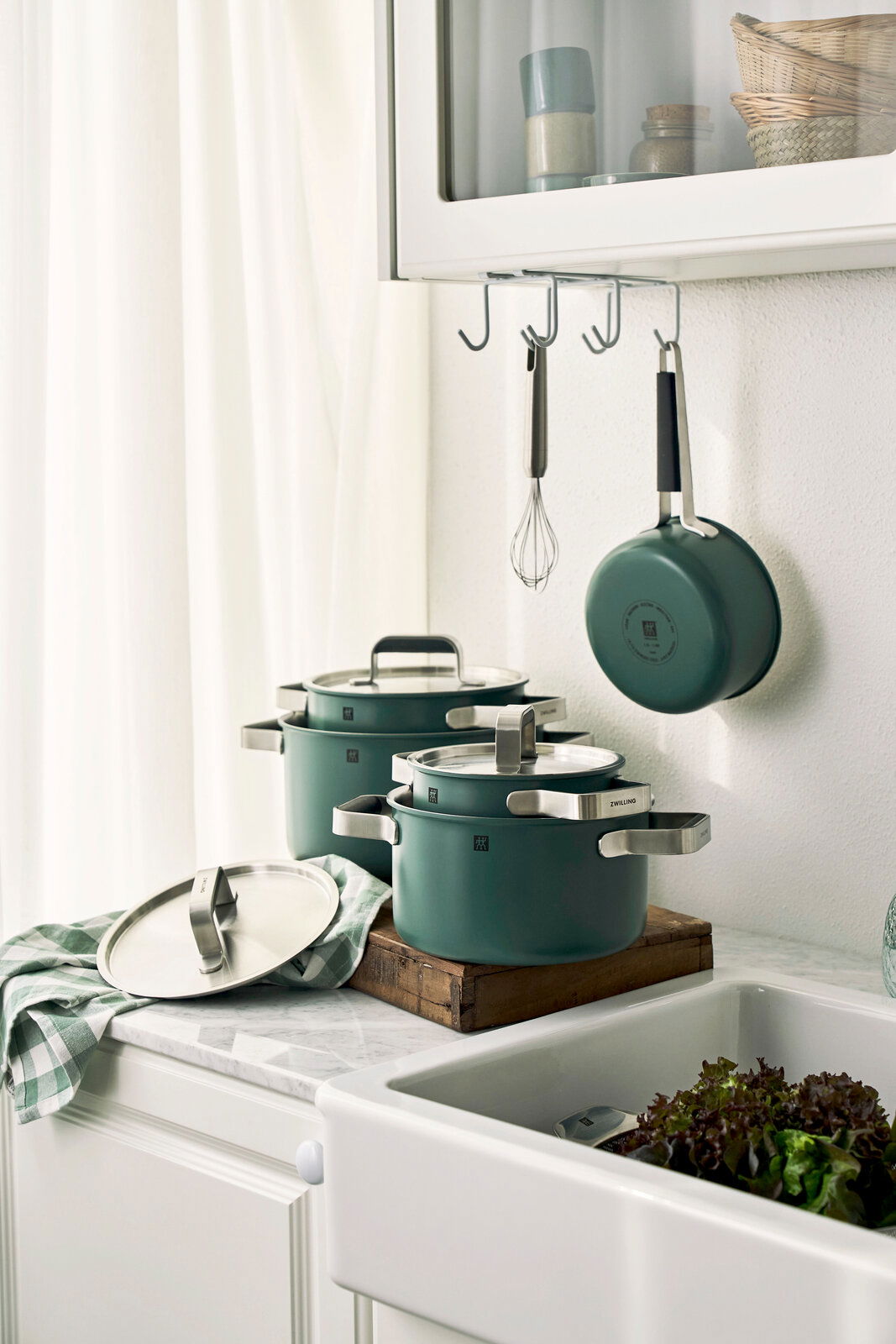 Küchenansicht mit einem 5-teiligen Topfset in modernem Design, bestehend aus grünen Töpfen mit Glasdeckeln und Edelstahlgriffen, auf einer Arbeitsplatte neben einer Spüle, mit Blick von der Seite.