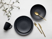 Dunkelblaues/anthrazitfarbenes Geschirrset mit geriffelter Strahlenstruktur (Teller und Schale) auf weißem Hintergrund, dazu goldfarbenes Besteck und schwarze Tasse; Draufsicht (Flatlay)