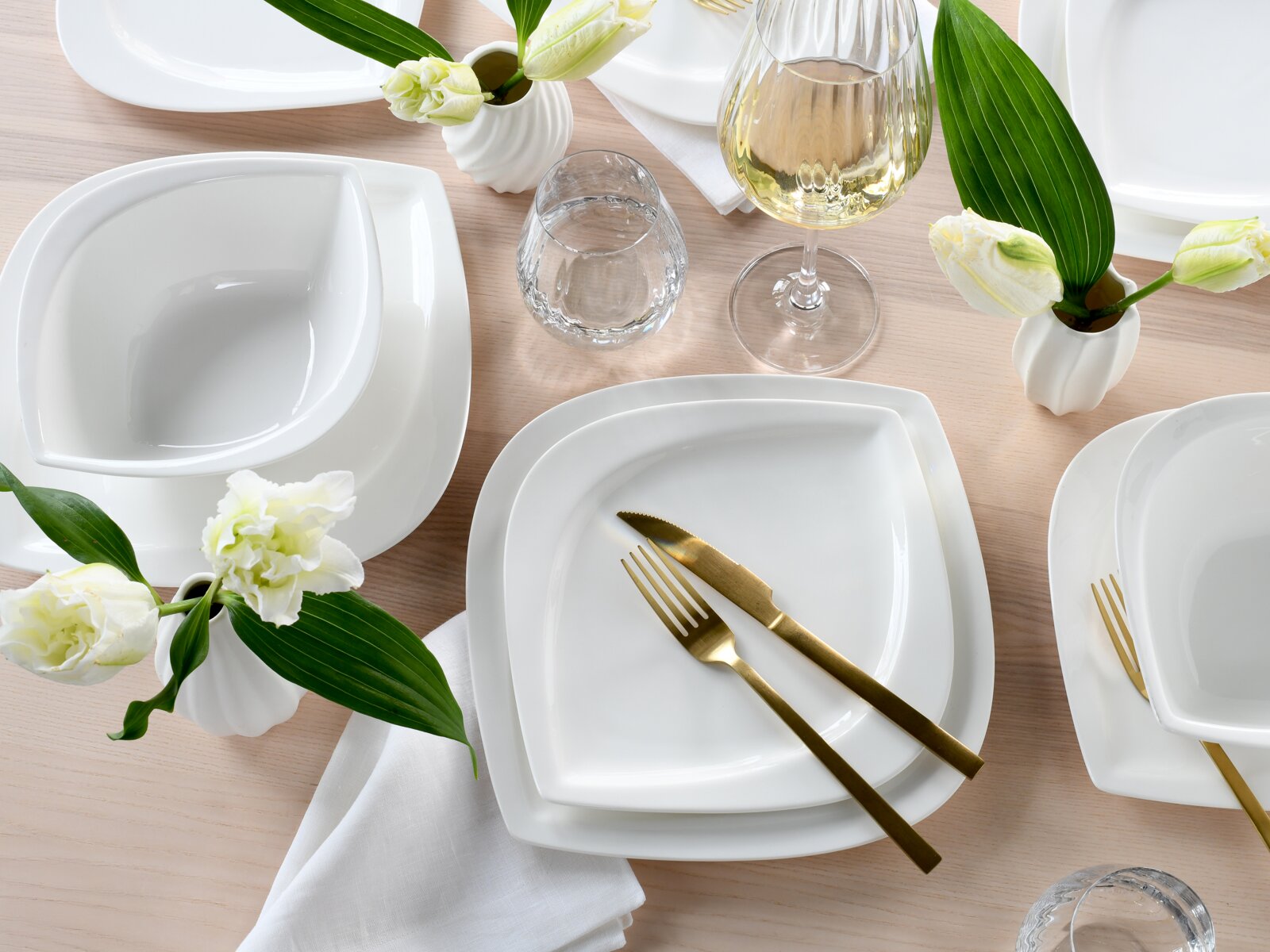 Draufsicht auf elegant gedeckten Tisch mit weißem, quadratisch abgerundetem Porzellan-Geschirrset, goldfarbenem Besteck, Wasser- und Weinglas sowie kleinen weißen Blumenvasen auf hellem Holztisch.