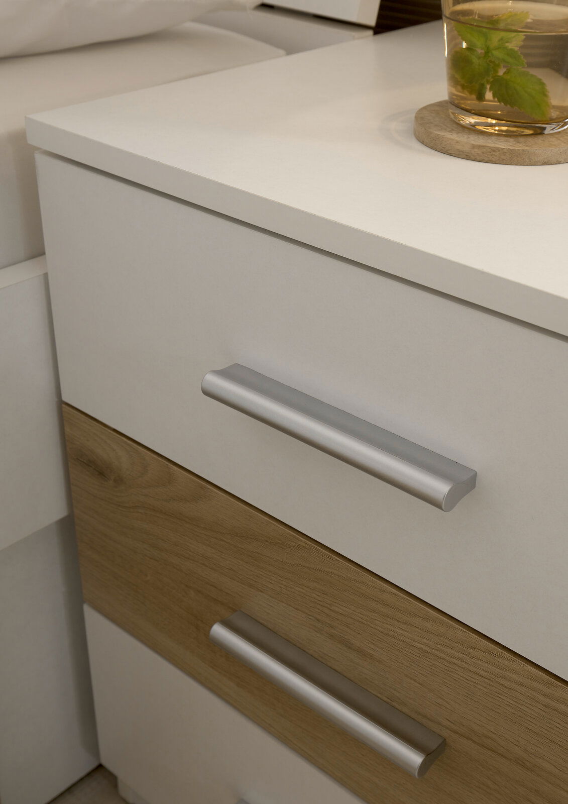 Nahaufnahme einer modernen Nachttischschublade mit weißer und holzfarbener Front, silbernen Griffen und einem Glas Tee auf der Oberfläche.
