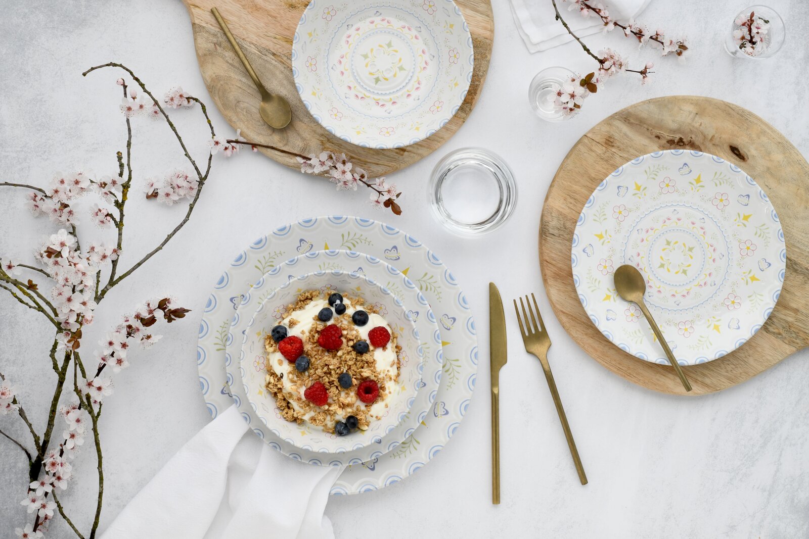 Draufsicht auf ein gedecktes Tisch-Setting mit weißem Porzellangeschirr im zarten Blumenmuster, goldfarbenem Besteck, Holzplatten, Wassergläsern und einer Müslischale mit Joghurt, Granola, Himbeeren und Blaubeeren, dekoriert mit blühenden Zweigen.