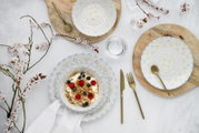 Draufsicht auf ein gedecktes Tisch-Setting mit weißem Porzellangeschirr im zarten Blumenmuster, goldfarbenem Besteck, Holzplatten, Wassergläsern und einer Müslischale mit Joghurt, Granola, Himbeeren und Blaubeeren, dekoriert mit blühenden Zweigen.