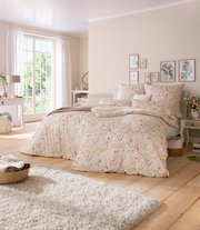 Gemütliches Schlafzimmer mit floraler Bettwäsche in Pastelltönen, Tageslicht durch großes Fenster, seitliche Perspektive