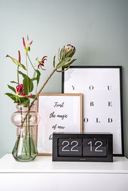 Tischuhr XL in modernem Design, frontal auf einem weißen Regal platziert, umgeben von dekorativen Elementen wie einer Vase mit Blumen und gerahmten Postern.