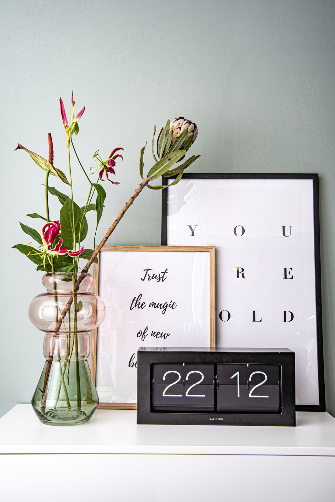 Tischuhr XL in modernem Design, frontal auf einem weißen Regal platziert, umgeben von dekorativen Elementen wie einer Vase mit Blumen und gerahmten Postern.