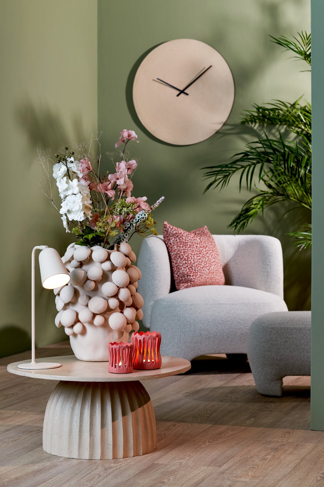Sfeervol modern interieur vanuit vooraanzicht, met een organisch gevormde fauteuil, een ronde salontafel, een decoratieve bloemenvaas, roze kaarsenhouders en een minimalistische wandklok tegen een saliegroene muur.