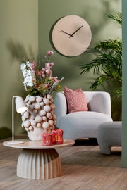 Sfeervol modern interieur vanuit vooraanzicht, met een organisch gevormde fauteuil, een ronde salontafel, een decoratieve bloemenvaas, roze kaarsenhouders en een minimalistische wandklok tegen een saliegroene muur.
