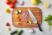 Vogelperspektive auf ein Schneidebrett mit einem Messer und geschnittenem Gemüse, umgeben von frischen Zutaten wie Tomaten, Zucchini, Zwiebeln und Basilikum auf einem Marmortisch.