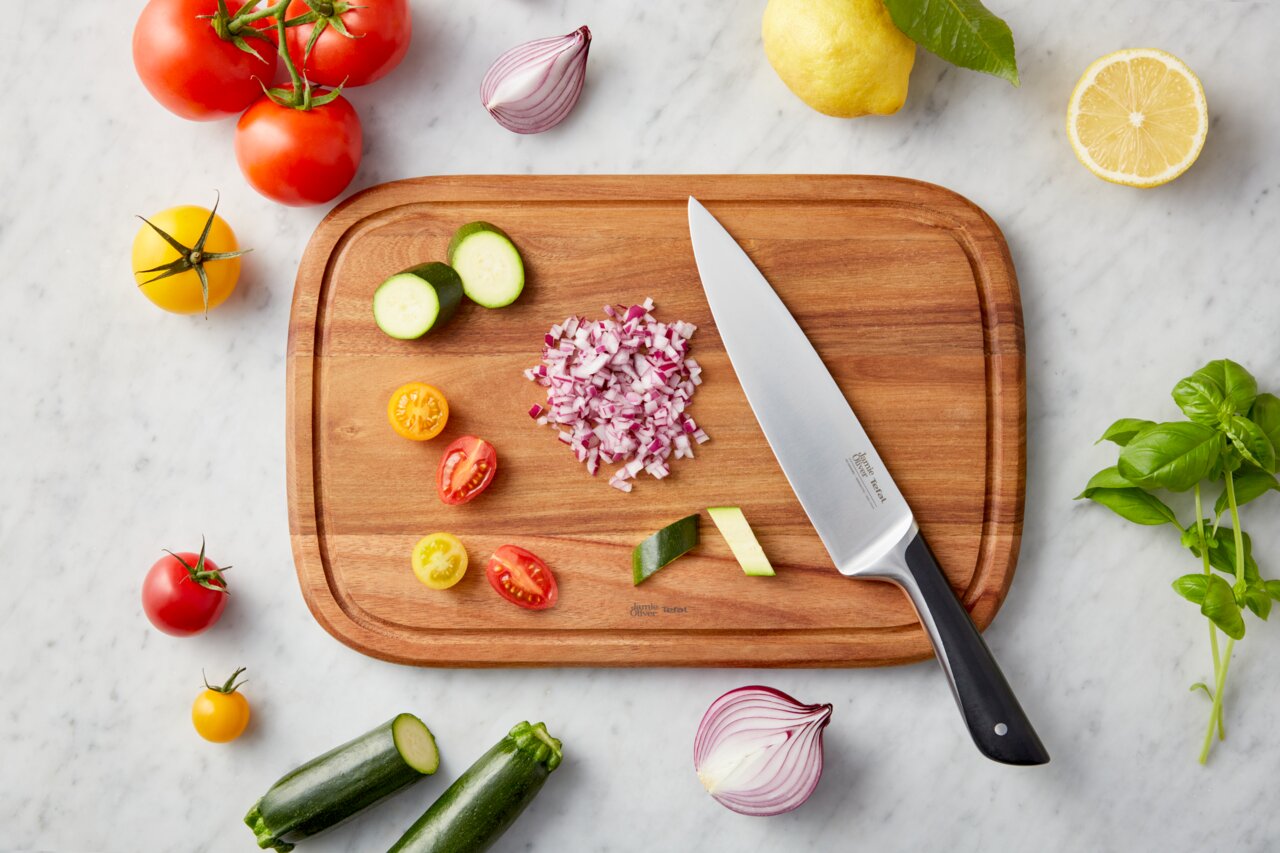 Vogelperspektive auf ein Schneidebrett mit einem Messer und geschnittenem Gemüse, umgeben von frischen Zutaten wie Tomaten, Zucchini, Zwiebeln und Basilikum auf einem Marmortisch.