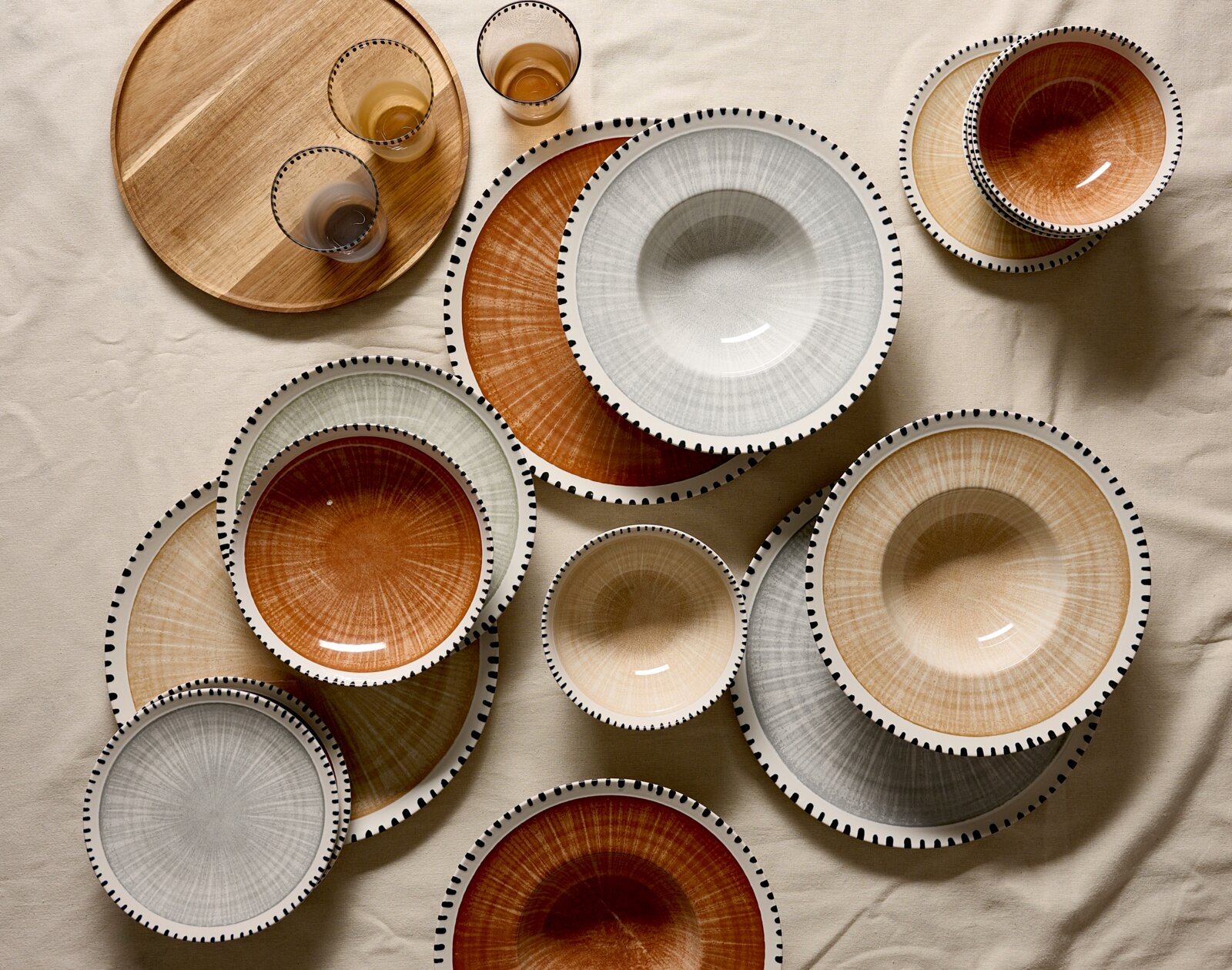 Bovenaanzicht van stijlvol servies met borden, kommen en glazen in warme aardetinten en grijs, afgewerkt met een decoratieve zwarte stippelrand, naast een ronde houten serveerplank.