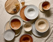 Bovenaanzicht van stijlvol servies met borden, kommen en glazen in warme aardetinten en grijs, afgewerkt met een decoratieve zwarte stippelrand, naast een ronde houten serveerplank.