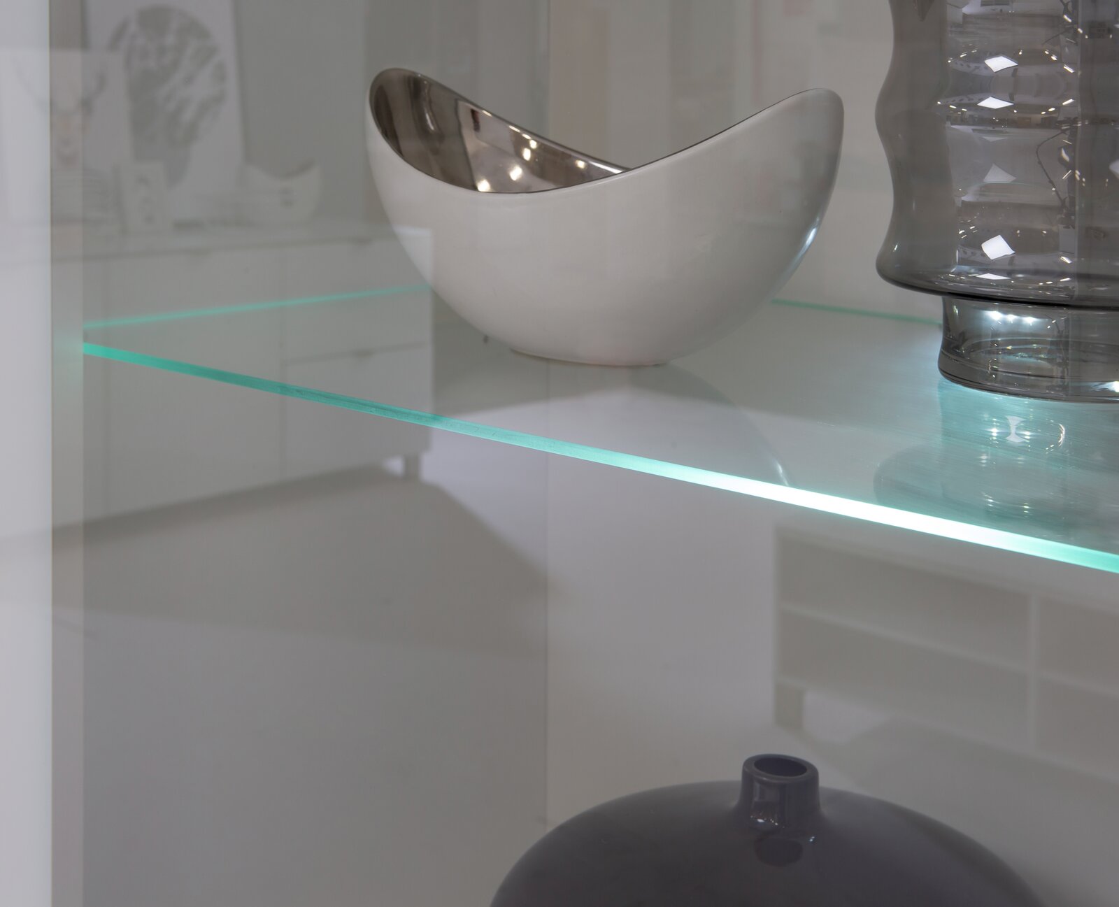 Nahaufnahme einer Vitrine mit Glasregal, auf dem eine silberne Schale und eine graue Vase stehen.