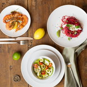 Vogelperspektive auf ein Geschirrset mit drei Tellern, die mit Garnelen, Salat und Pasta angerichtet sind, auf einem Holztisch.