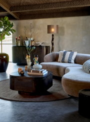 Modernes Wohnzimmer aus frontaler Perspektive mit geschwungenem taupefarbenem Sofa, dunklem Couchtisch, Sideboard und dekorativer Stehleuchte