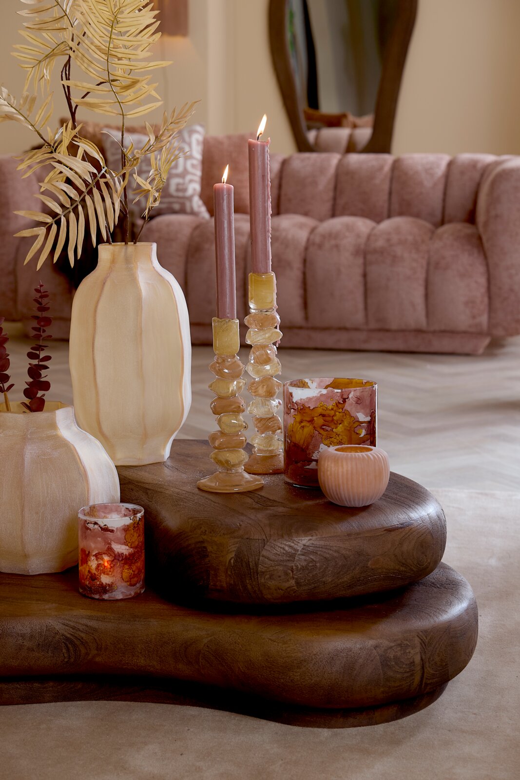 Stimmungsvolles Wohnaccessoire-Arrangement mit rosafarbenen Stabkerzen in organisch geformten Glashaltern, dekorativen Windlichtern und beigefarbenen Vasen auf einem massiven Holztisch, aus leicht erhöhter Frontalperspektive inszeniert.