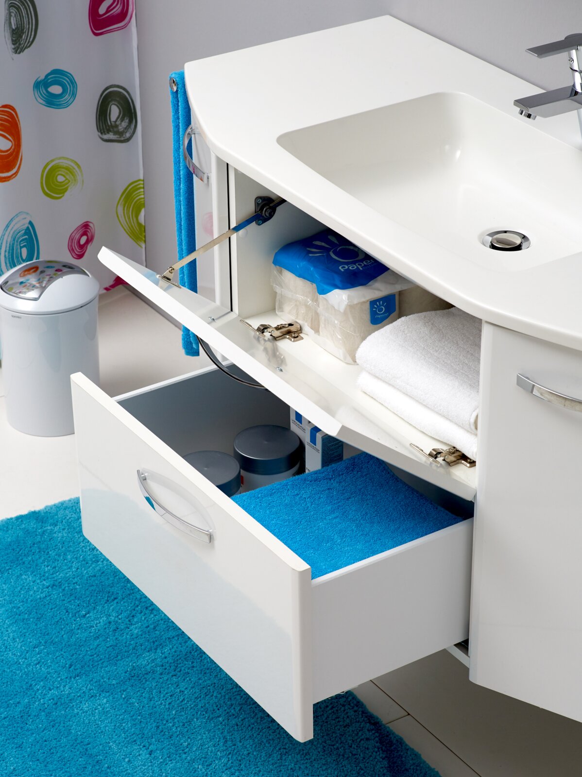 Weißer Waschtischunterschrank im Badezimmer mit integriertem Waschbecken, halb geöffneter Klappe und ausgezogenem Schubfach zur Aufbewahrung von Handtüchern und Pflegeartikeln, Perspektive: schräg von vorn rechts.