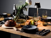 Stilvoll gedeckter Holztisch mit schwarzem Geschirr, geriffelten Rauchglas-Gläsern, Weingläsern, moderner Tischlampe, großer Glasvase mit weißen Blumen, Brot und Birnen, aus leicht schräger Perspektive von vorne auf Tischhöhe aufgenommen.