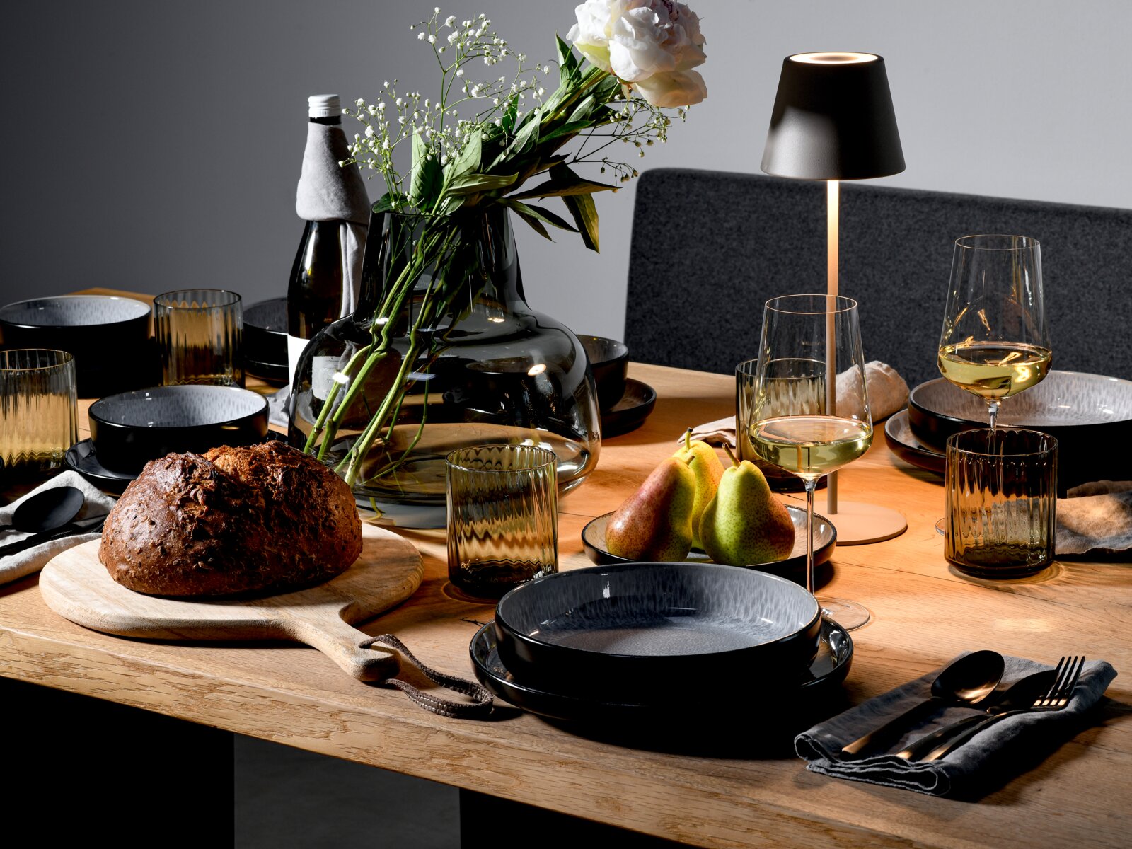 Stilvoll gedeckter Holztisch mit schwarzem Geschirr, geriffelten Rauchglas-Gläsern, Weingläsern, moderner Tischlampe, großer Glasvase mit weißen Blumen, Brot und Birnen, aus leicht schräger Perspektive von vorne auf Tischhöhe aufgenommen.