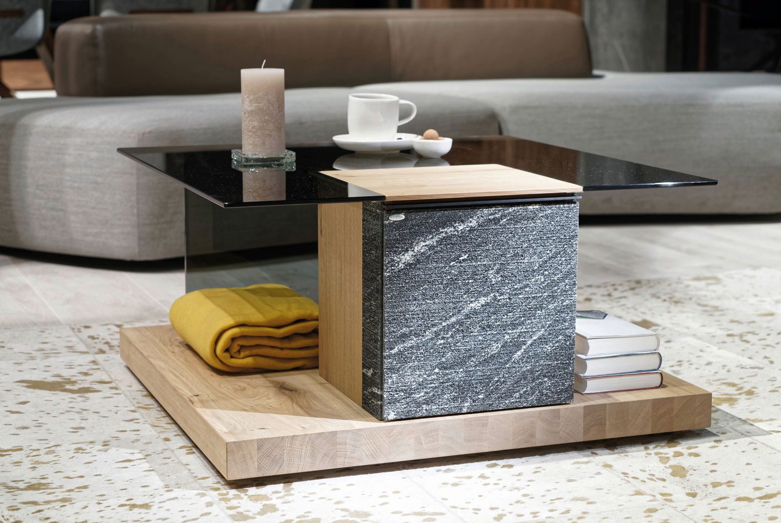 Moderner Couchtisch mit schwarzer Glasplatte, Holz- und Steinelementen, von der Seite fotografiert. Dekoriert mit einer Kerze, einer Tasse und Büchern.