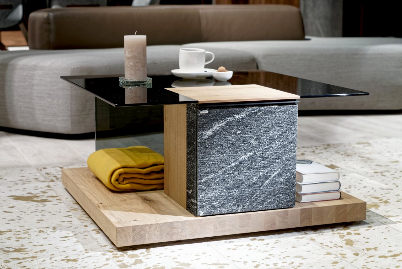 Moderner Couchtisch mit schwarzer Glasplatte, Holz- und Steinelementen, von der Seite fotografiert. Dekoriert mit einer Kerze, einer Tasse und Büchern.