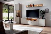 Modernes Wohnzimmer mit hellgrauer TV-Wohnwand und Holz-Akzenten, Lowboard mit großem Fernseher, Vitrine mit Beleuchtung und Wandregal; perspektivisch aus leicht schräger Frontansicht aufgenommen.