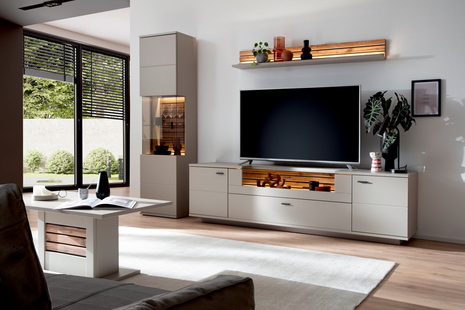Modernes Wohnzimmer mit hellgrauer TV-Wohnwand und Holz-Akzenten, Lowboard mit großem Fernseher, Vitrine mit Beleuchtung und Wandregal; perspektivisch aus leicht schräger Frontansicht aufgenommen.