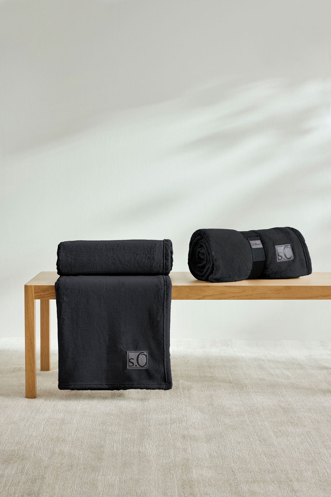 Zwei schwarze s.Oliver Kuschelsoft-Decken auf einer Holzbank, eine gefaltet und eine gerollt, in einem hellen Raum.