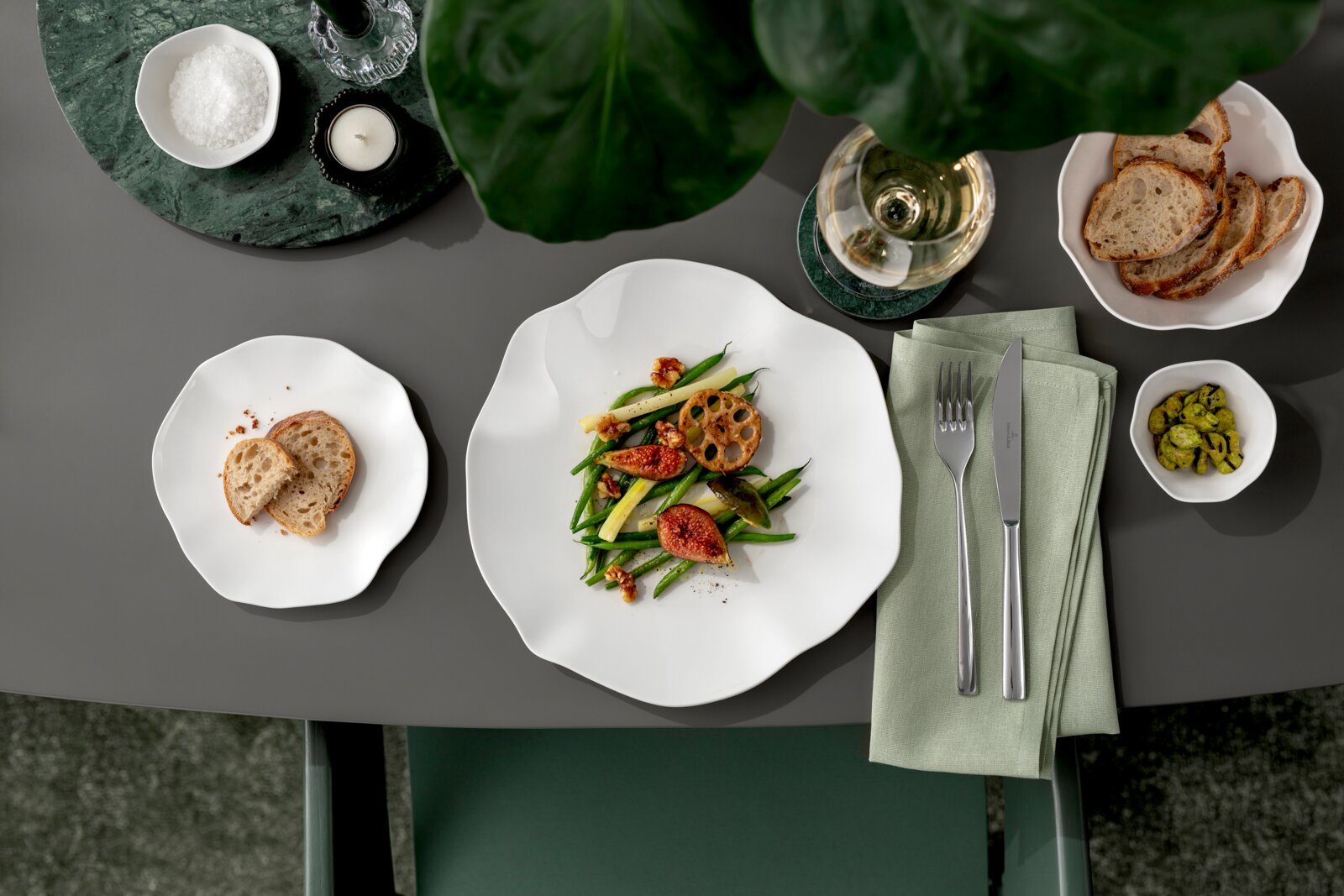 Draufsicht auf einen gedeckten Tisch mit weißen, organisch geformten Porzellantellern: Hauptteller mit grünem Gemüse und Feigen, Brotscheiben, Schale mit Pistazien, Glas Weißwein, Besteck auf grünem Stoffserviettenstapel sowie Kerze und Salzschale.