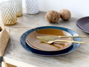 Schräg von oben fotografiertes Tisch-Setting mit blauem Speiseteller und braun glasierter Schale, darauf goldfarbenes Messer und Gabel auf rustikalem Holztisch, dekoriert mit Kerzenhaltern und Naturdeko.