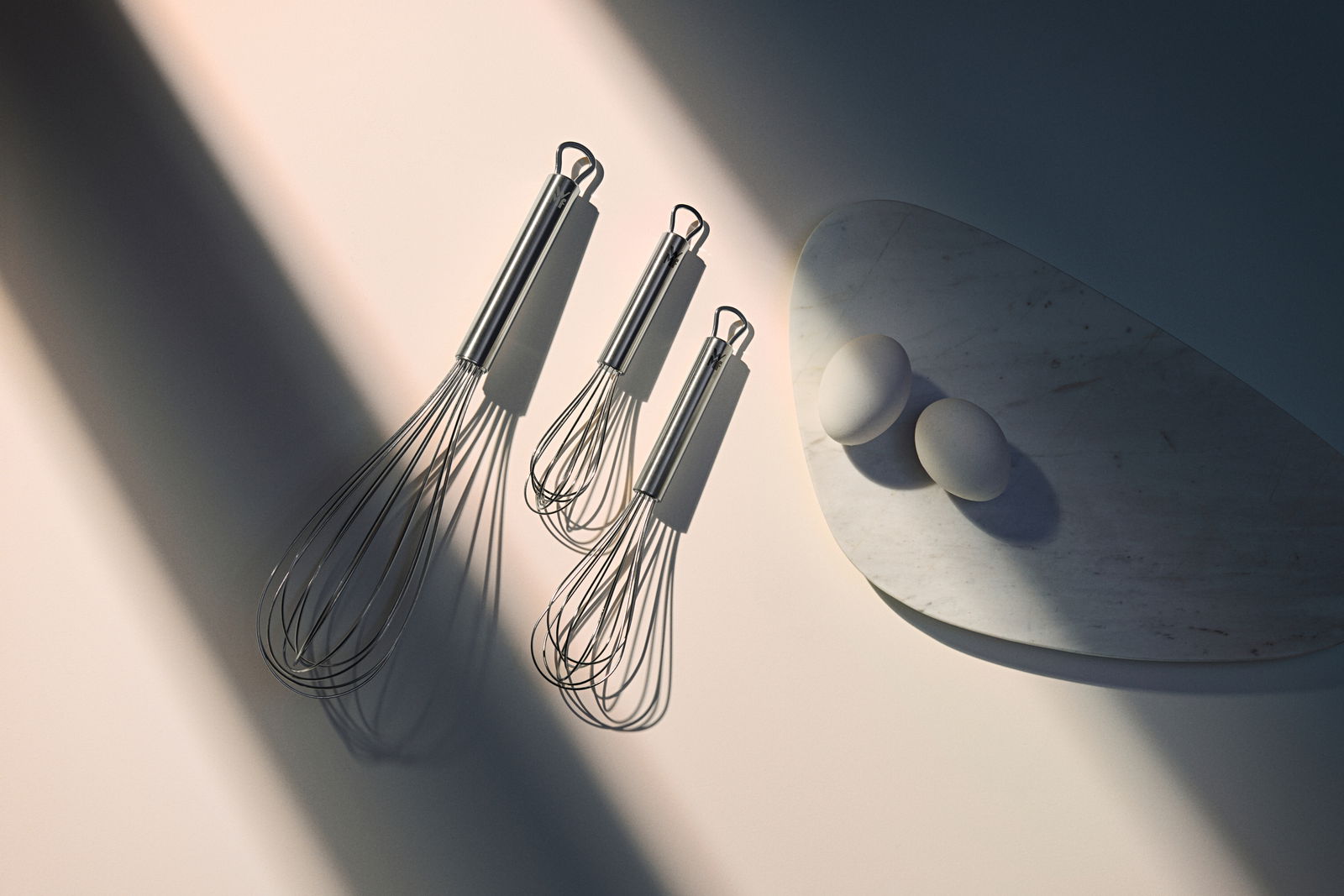 WMF Schneebesen 3-tlg. PROFI PLUS Drei Schneebesen aus Edelstahl in verschiedenen Größen, von oben betrachtet, neben zwei Eiern auf einem Marmorbrett.