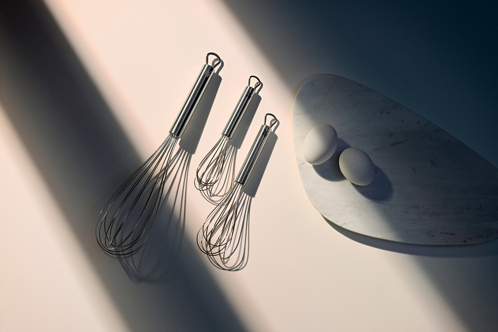 Drei Schneebesen aus Edelstahl in verschiedenen Größen, von oben betrachtet, neben zwei Eiern auf einem Marmorbrett.