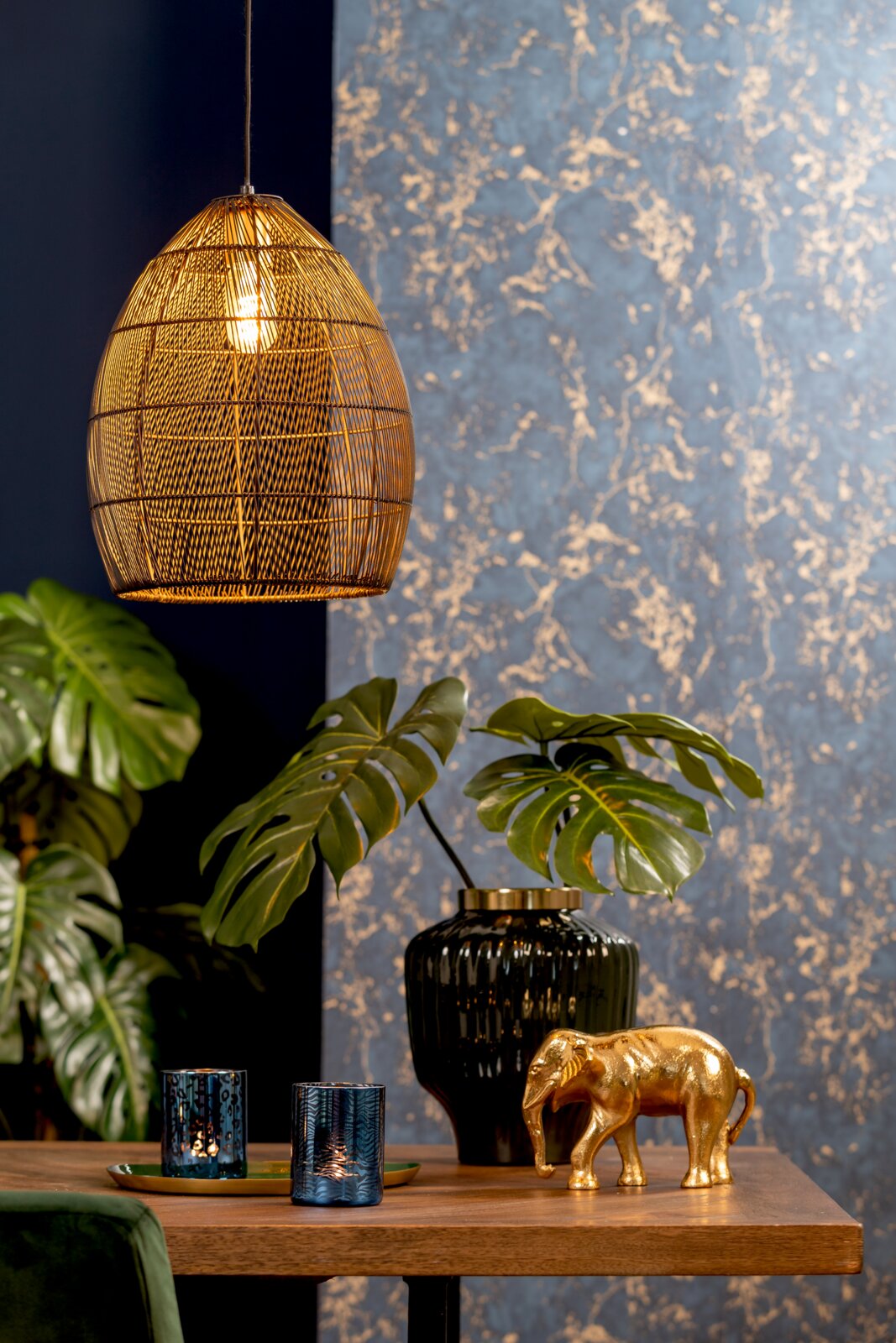 Schuin vooraanzicht van een sfeervolle goudkleurige gevlochten hanglamp boven een houten tafel met decoratieve accessoires en groene planten.