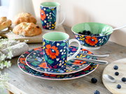 Buntes Geschirr-Set mit floralem Muster: Tasse auf passenden Tellern, Schale mit Blaubeeren und weitere Tasse im Hintergrund, aufgenommen in leichter Seitenansicht auf einem Holztisch.