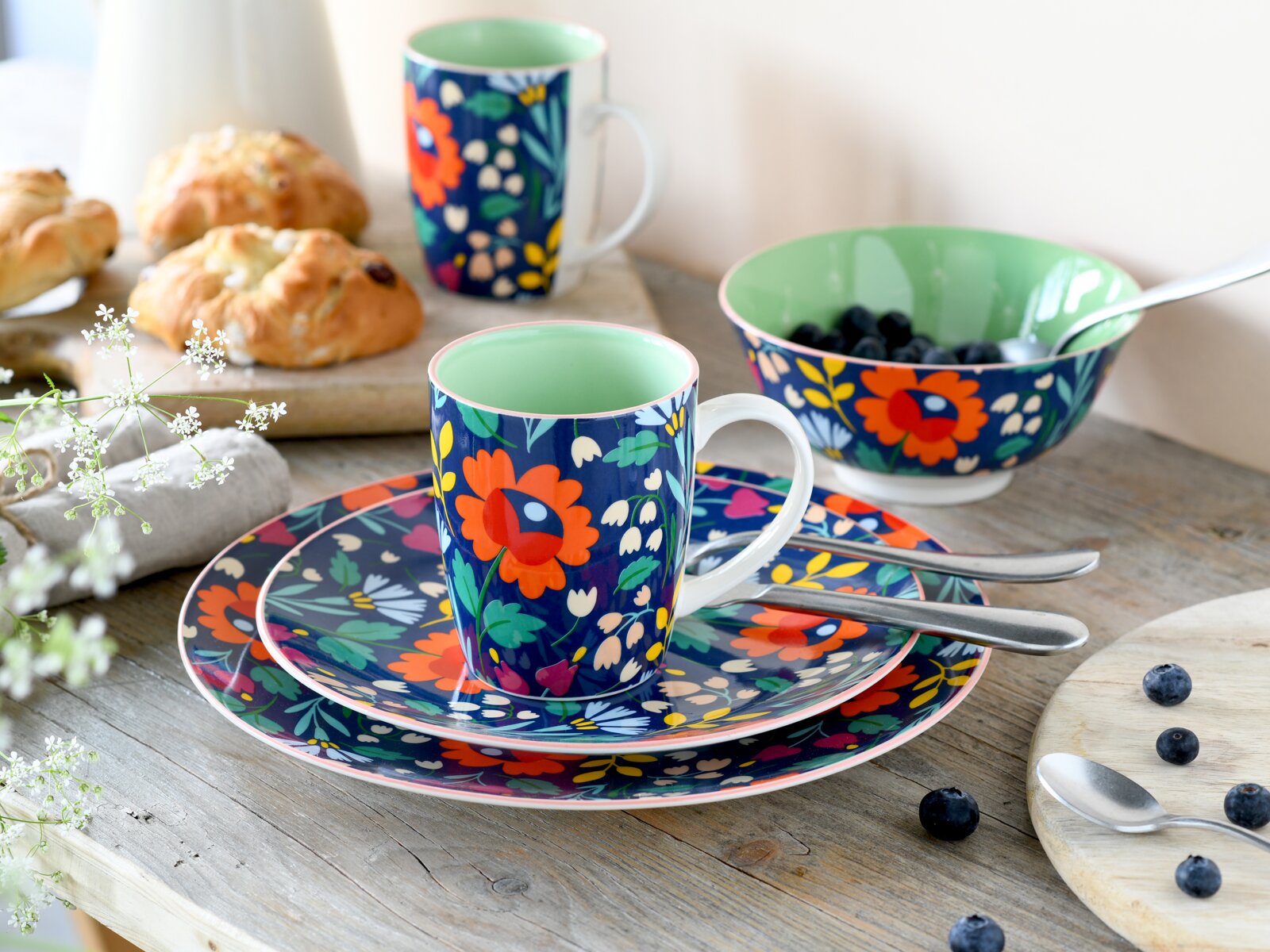 Buntes Geschirr-Set mit floralem Muster: Tasse auf passenden Tellern, Schale mit Blaubeeren und weitere Tasse im Hintergrund, aufgenommen in leichter Seitenansicht auf einem Holztisch.