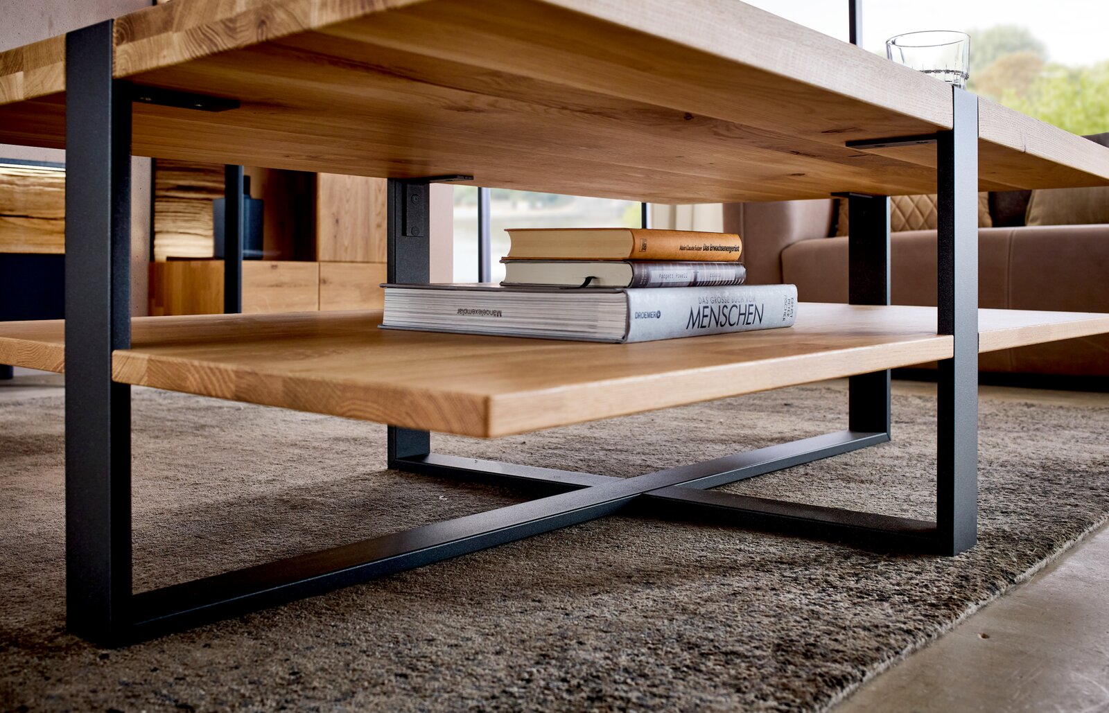 Moderner Couchtisch aus massiver Holzplatte mit schwarzem Metallgestell und Ablagefach, mit Büchern dekoriert, in bodennaher Seitenansicht aus leichtem Untersichtwinkel im Wohnzimmer auf Teppich.