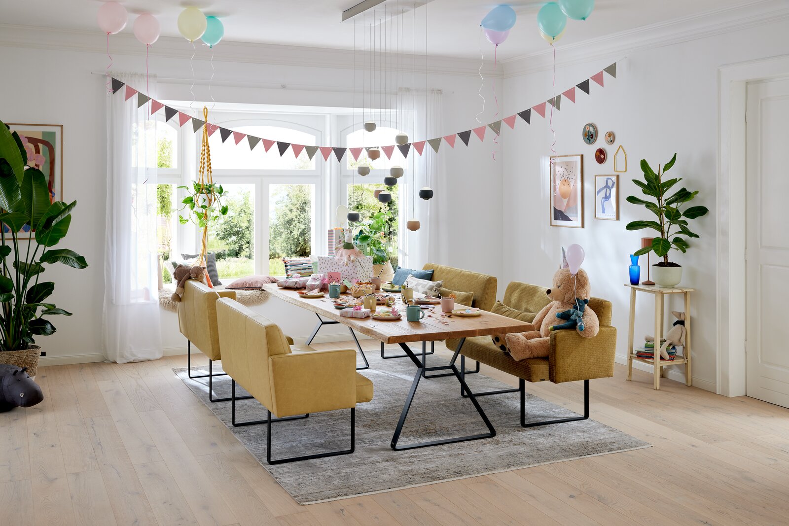 Gemütliche Esszimmer-Szene mit gelben Bänken und einem Holztisch, dekoriert für eine Feier mit Luftballons und Girlanden. Perspektive von der Seite.