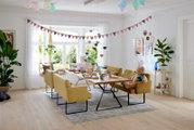 Gemütliche Esszimmer-Szene mit gelben Bänken und einem Holztisch, dekoriert für eine Feier mit Luftballons und Girlanden. Perspektive von der Seite.