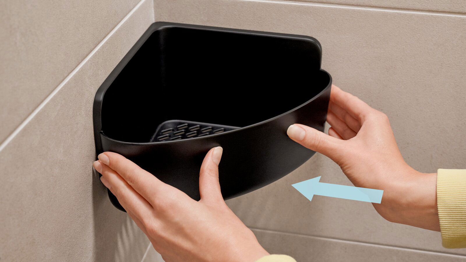 Schwarzer Eckduschkorb von tesa, der in einer Ecke an der Wand montiert wird. Perspektive von vorne mit Händen, die den Korb halten.