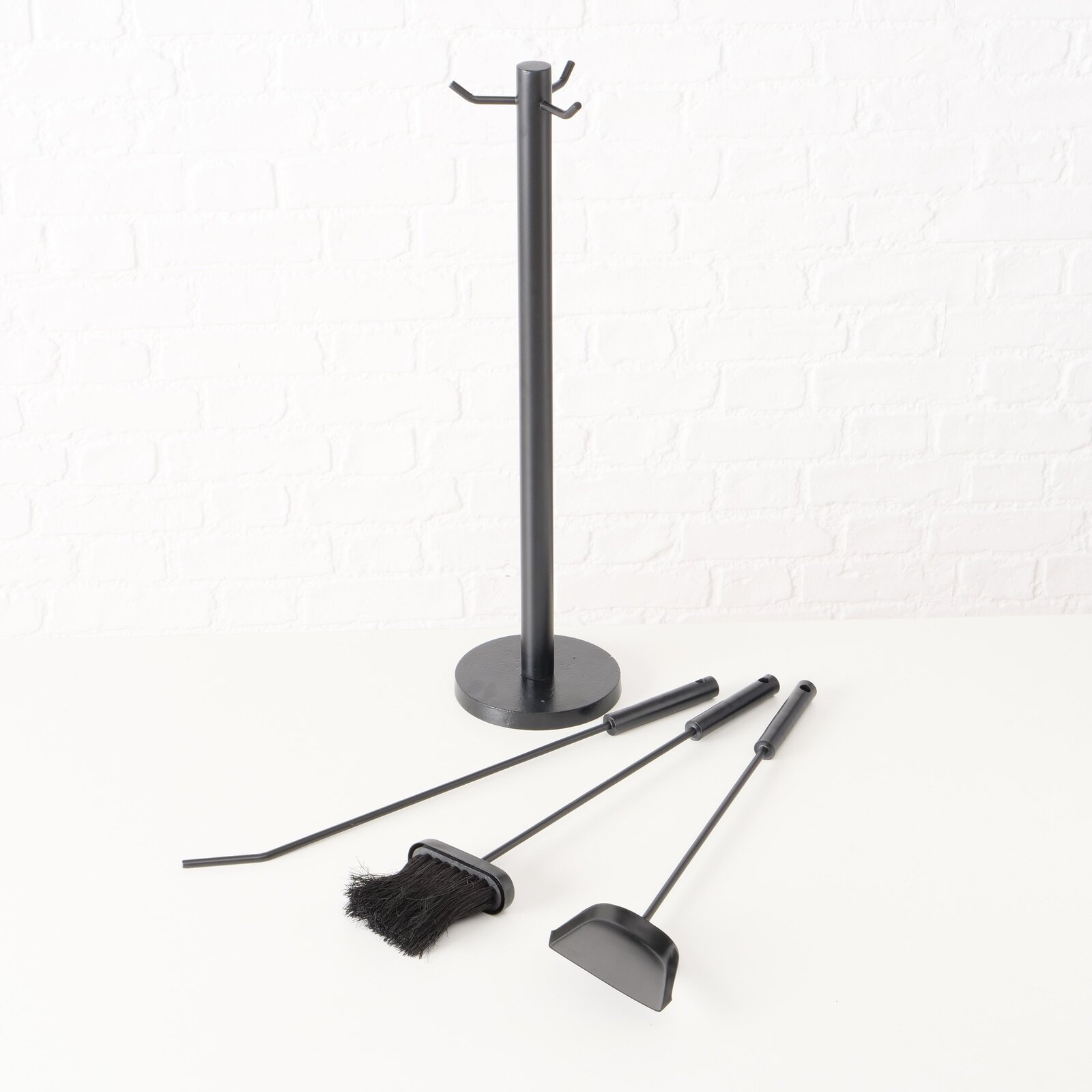 Kaminbesteck Mika, 4-teilig, bestehend aus einem Ständer und drei Werkzeugen, auf einer weißen Oberfläche vor einer weißen Ziegelwand, seitliche Perspektive.