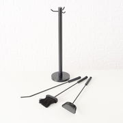 Kaminbesteck Mika, 4-teilig, bestehend aus einem Ständer und drei Werkzeugen, auf einer weißen Oberfläche vor einer weißen Ziegelwand, seitliche Perspektive.